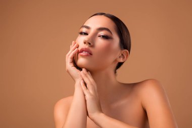 A stylish brunette woman poses gracefully against a beige backdrop, showcasing smooth skin, well-applied makeup, and a sophisticated expression, embodying modern beauty and elegance.