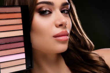 Close-up of a young woman with professional makeup holding a cosmetics palette. Highlighting beauty, fashion, and skincare themes with a sophisticated and stylish ambiance, set against a dark green backdrop.