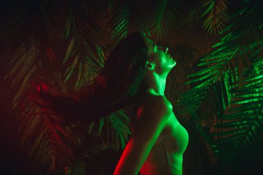 Beautiful young woman stands in tropical jungle setting lit with green and red neon lights, creating an exotic and striking aesthetic.