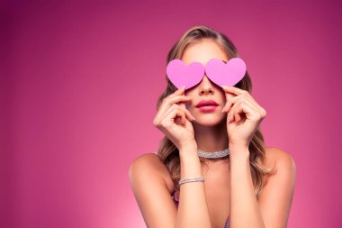 Elegant young blonde woman concealing her eyes with heart shapes, styled beautifully in jewelry and makeup, set against a vibrant pink background with a sense of glamour and sophistication.