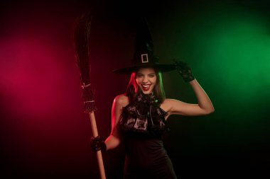 Beautiful witch wearing fashionable black outfit and hat, posing with broom against vibrant dark background, perfect for Halloween celebration visuals and magical fantasy scenes