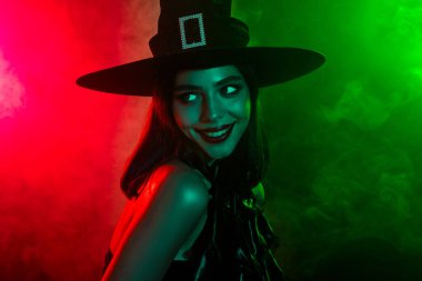 A mystifying woman dressed as a witch in a dark outfit and hat, with red and green atmospheric lighting exuding a magical Halloween ambiance.