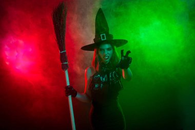 A captivating witch in a dark costume stands with her broomstick amidst red and green fogs, embodying mystique and enchantment perfect for a Halloween scene.