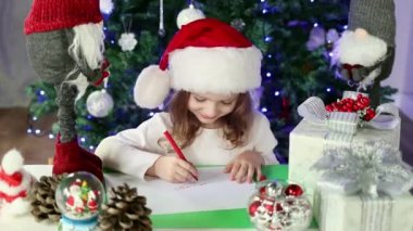 Kırmızı Noel Baba şapkalı küçük bir kız Noel Baba 'ya Noel ağacının altına dilekleriyle bir mektup yazıyor. Noel ve Yeni Yıl atmosferi.
