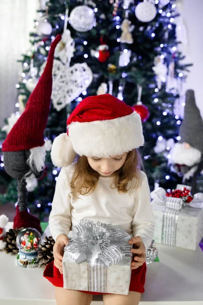 Kırmızı Noel Baba şapkalı küçük bir kız Noel ağacının altında oturuyor ve hediye kutusuyla oynuyor. Noel ve Yeni Yıl atmosferi. Mutlu bir çocukluk kavramı.