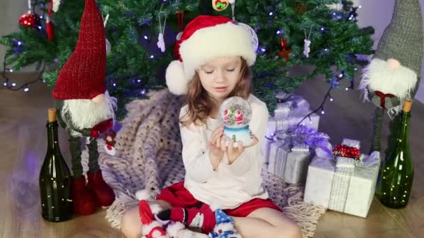 一个穿着红色圣诞老人帽的小女孩正坐在圣诞树下玩着一个神奇的雪球 圣诞和新年气氛 幸福童年的概念 慢动作 — 图库视频影像