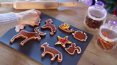 Evde Noel kurabiyelerini yavaş çekimde boyayan kadınların elleri. Mutfakta Noel süsü var. Peri ışıklarıyla fidan ağacı. Yeni yıl ve Noel konsepti. Tatil atmosferi.