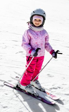3 yaşında, Avrupa görünümlü pembe tulumlu, kar dağlarında kayak yaparken gülümseyen küçük bir kız. Kış aktivitesi. Dinlenmek, hobiler, sağlık.
