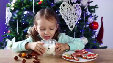 A little curly-haired girl drinks milk from a glass, eats gingerbread Christmas cookies, and laughs happily against a background of a Christmas tree. The atmosphere of Christmas and New Year.