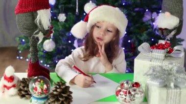 Kırmızı Noel Baba şapkalı küçük bir kız Noel Baba 'ya Noel ağacının altına dilekleriyle bir mektup yazıyor. Noel ve Yeni Yıl atmosferi. Yavaş çekim.