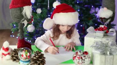 Kırmızı Noel Baba şapkalı küçük bir kız Noel Baba 'ya Noel ağacının altına dilekleriyle bir mektup yazıyor. Noel ve Yeni Yıl atmosferi. Yavaş çekim.