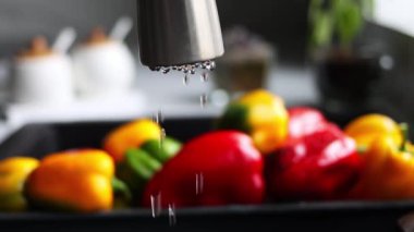 Musluktan damlayan su damlaları yavaş çekimde kırmızı ve sarı biberlerin arka planına doğru akar. Su akıntısı lavabodaki biberin üzerine dökülüyor. Modern bir mutfağın içi.