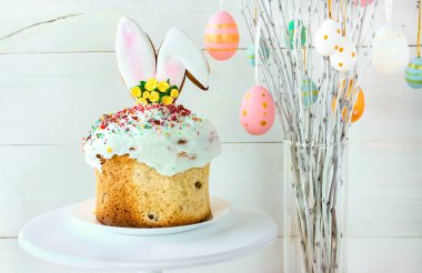 Easter cake stands on a white plate on a white wooden background. The branches of the trees decorated with multi-colored Easter eggs stand in a vase. Minimal Easter composition with place for text.