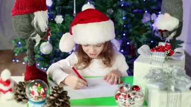 Kırmızı Noel Baba şapkalı küçük bir kız Noel Baba 'ya Noel ağacının altına dilekleriyle bir mektup yazıyor. Noel ve Yeni Yıl atmosferi.