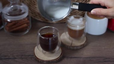 Bir kadının eli, bir Türk köpüğüyle sütü cam bardaktaki taze kahveye döker. Kahvaltı kahve konsepti. Evde kahve yapma sürecinde. Yavaş çekim.