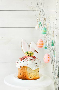 Easter cake stands on a white plate on a white wooden background. The branches of the trees decorated with multi-colored Easter eggs stand in a vase. Minimal Easter composition with place for text.