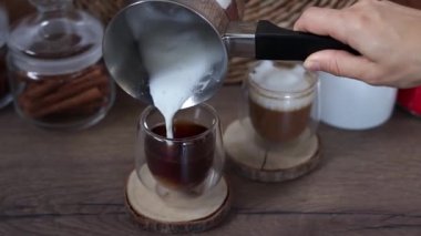 Bir kadının eli, bir Türk köpüğüyle sütü cam bardaktaki taze kahveye döker. Kahvaltı kahve konsepti. Evde kahve yapma sürecinde. Yavaş çekim.