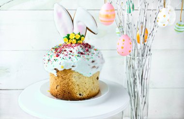 Easter cake stands on a white plate on a white wooden background. The branches of the trees decorated with multi-colored Easter eggs stand in a vase. Minimal Easter composition with place for text.