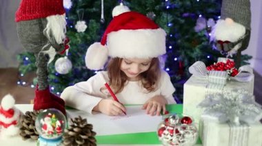 Kırmızı Noel Baba şapkalı küçük bir kız Noel Baba 'ya Noel ağacının altına dilekleriyle bir mektup yazıyor. Noel ve Yeni Yıl atmosferi. Yavaş çekim.
