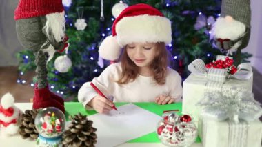 Kırmızı Noel Baba şapkalı küçük bir kız Noel Baba 'ya Noel ağacının altına dilekleriyle bir mektup yazıyor. Noel ve Yeni Yıl atmosferi. Yavaş çekim.