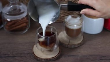 Bir kadının eli, bir Türk köpüğüyle sütü cam bardaktaki taze kahveye döker. Kahvaltı kahve konsepti. Evde kahve yapma süreci..