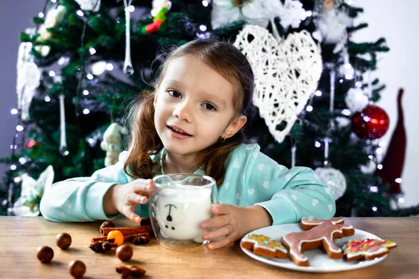 Kıvırcık saçlı küçük bir kız bardaktan süt içer ve bir Noel ağacının arka planında mutlu bir şekilde güler. Noel ve Yeni Yıl atmosferi.