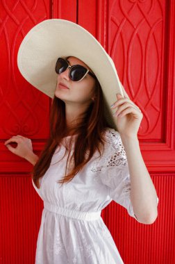 Şapkalı, beyaz elbiseli ve gözlüklü çekici bir genç kız eski kırmızı bir kapının yanında poz veriyor..