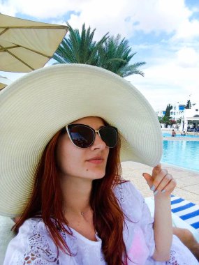 Şapkalı çekici genç bir kız yüzme havuzunun yanında oturuyor ve gülümsüyor. Turizm, tatil ve dinlenme konsepti. Otel kompleksinin reklamı..