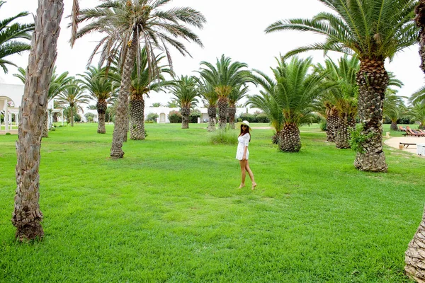 ヤシの木の下の緑の芝生の上に帽子の中にヨーロッパの外観の若いかなりの女の子が立っています 熱帯の島 観光とレクリエーションの概念 — ストック写真
