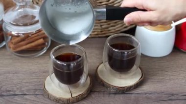Bir kadının eli, bir Türk köpüğüyle sütü cam bardaktaki taze kahveye döker. Kahvaltı kahve konsepti. Evde kahve yapma süreci..