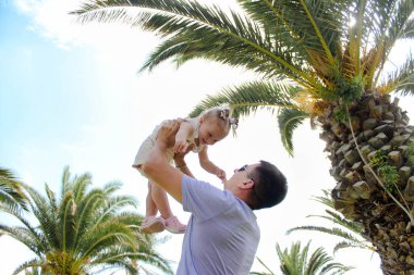 Genç bir baba, küçük kızını arka plandaki palmiye ağaçlarına yaslar. Mutlu çocukluk, ebeveynlik ve mutlu aile kavramı. Turizm ve eğlence kavramı.