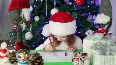 Kırmızı Noel Baba şapkalı küçük bir kız Noel Baba 'ya Noel ağacının altına dilekleriyle bir mektup yazıyor. Noel ve Yeni Yıl atmosferi. Yavaş çekim.