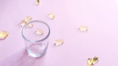 Vitaminli şeffaf sarı kapsüller pembe arka planda duran şeffaf bir bardağa dökülüyor. Balık yağı takviyesi. Sağlık, vitamin ve ilaç kavramı. Omega 3, D vitamini. 4K görüntüsü.
