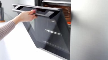 The woman's hands open the oven and pull a baking sheet out of the oven. A young woman in a white modern kitchen cooking food in an oven close up. Concept of Christmas and New Year.