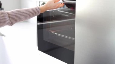 The womans hands open the oven and stick the baking sheet with ginger biscuits there. A young woman in a white modern kitchen cooking food in an oven close up. Concept of Christmas and New Year.