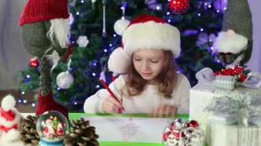 Kırmızı Noel Baba şapkalı küçük bir kız Noel Baba 'ya Noel ağacının altına dilekleriyle bir mektup yazıyor. Noel ve Yeni Yıl atmosferi. Yavaş çekim.