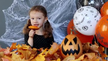 A little witch girl is funny eating a slice of pumpkin on the background of Halloween attributes in slow motion. Halloweens holiday attributes. Lantern carved from pumpkin known as Jack-o-lantern.
