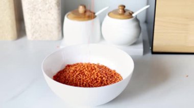 Kırmızı mercimek, modern bir mutfağın arka planına karşı ağır çekimde beyaz bir tabağa dökülür. Sağlıklı gıda ve beslenme konsepti. Güvenli, ekolojik ve sağlıklı ürünler..