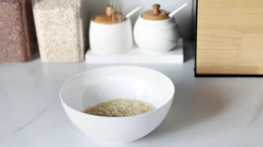 Beyaz çiğ pirinç, modern bir mutfağın arka planına karşı ağır çekimde beyaz bir tabağa dökülür. Sağlıklı gıda ve beslenme konsepti. Güvenli, ekolojik ve sağlıklı ürünler.