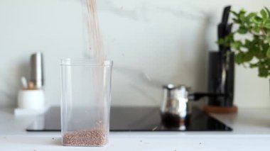 Karabuğday tarlaları, modern bir mutfağın arka planına karşı şeffaf bir kaba yavaş çekimde dökülür. Sağlıklı gıda ve beslenme konsepti. Güvenli, ekolojik ve sağlıklı ürünler