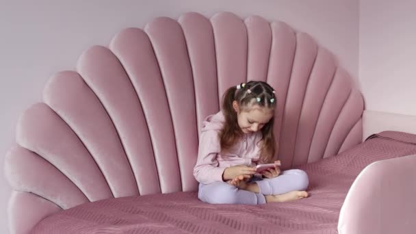 ヨーロッパ出身の少女がベッドの上に座り 携帯電話を使ってゆっくりと動いている 子供は携帯電話で仮想ゲームをしたり スマートフォンで漫画を見たりしています — ストック動画