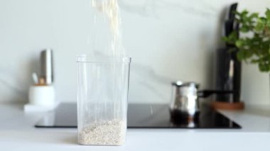 Yulaf ezmesi, modern bir mutfağın arka planına karşı şeffaf bir kaba ağır çekimde dökülür. Sağlıklı gıda ve beslenme konsepti. Güvenli, ekolojik ve sağlıklı ürünler. Ekolojik ambalaj, haşere kontrolü