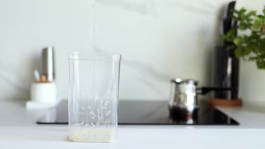 Beyaz çiğ pirinç, modern bir mutfağın arka planına karşı şeffaf bir kaba ağır çekimde dökülür. Sağlıklı gıda ve beslenme konsepti. Güvenli, ekolojik ve sağlıklı ürünler. Ekolojik