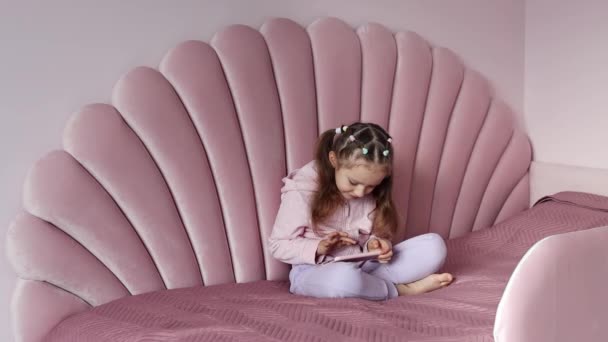 ヨーロッパ出身の少女がベッドの上に座り 携帯電話を使ってゆっくりと動いている 子供は携帯電話で仮想ゲームをしたり スマートフォンで漫画を見たりしています — ストック動画