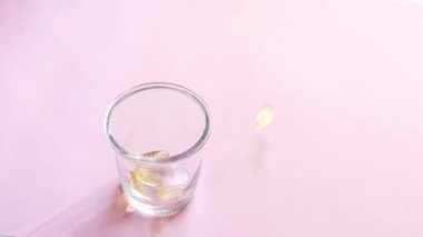 Vitaminli şeffaf sarı kapsüller pembe arka planda duran şeffaf bir bardağa dökülüyor. Balık yağı takviyesi. Sağlık, vitamin ve ilaç kavramı. Omega 3, D vitamini.