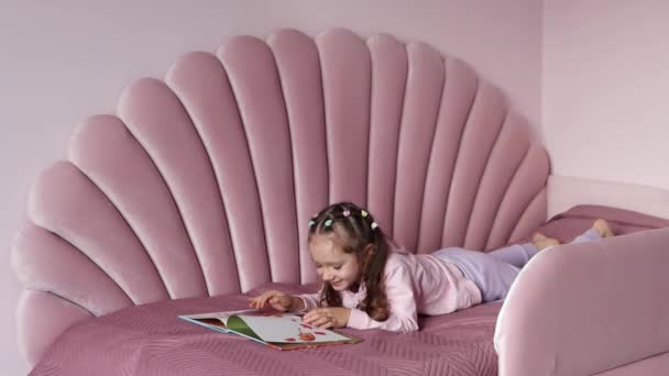 小さな女の子が本を見ながらベッドに横たわっている 本やスマートフォンを選択するには 子供時代とレジャーの概念 4K映像について — ストック動画