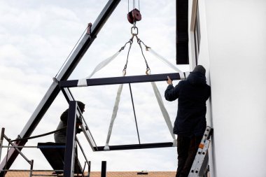 İnşaat alanında iki işçinin bir vinci var. Vinç metal yapıyı kaldırır. Kargo bir vinç tarafından havaya kaldırıldı..