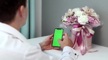 Erkek ellerinin bir buket çiçeğin yanında yeşil bir ekran telefon tutarken yakın plan çekimleri. Takım elbiseli genç bir adam internette Chroma tuşuna basıyor yazı yazarken elleri kapatıyor. Çevrimiçi kavramı