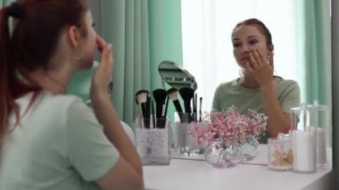 Genç bir kadın aynaya yavaş çekimde bakarken yüzüne krem sürüyor. Cilt bakımı. Güzellik anlayışı, kişisel bakım ve günlük krem kullanımı.