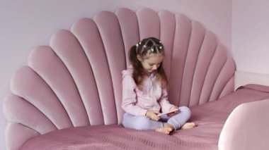 Avrupalı görünümlü küçük bir kız yatakta oturuyor ve cep telefonunu yavaş çekimde kullanıyor. Çocuk ya cep telefonuyla sanal oyunlar oynuyor ya da akıllı telefonuyla çizgi film izliyor. Çocuk.